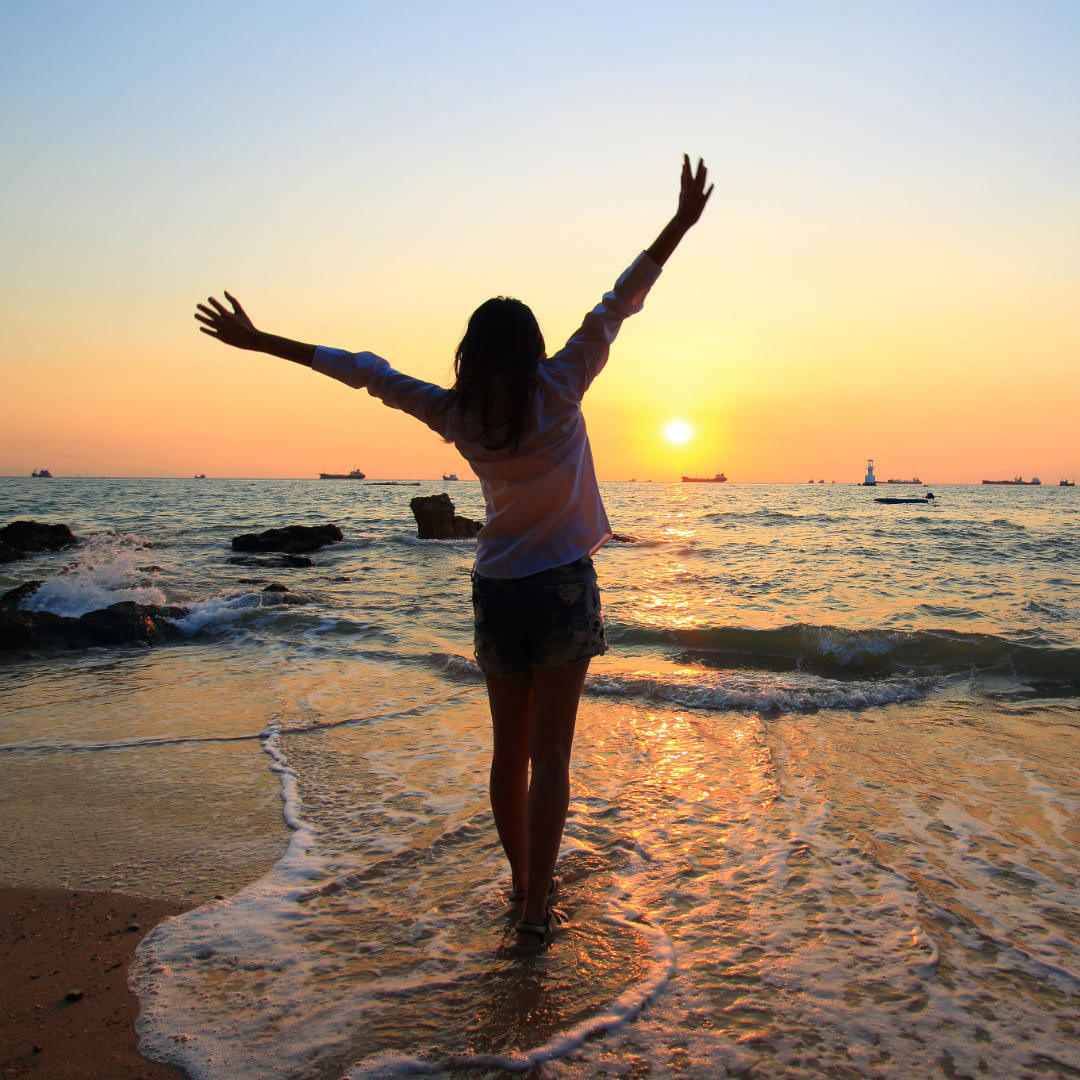 A woman stands on the beach with a gentle wave washing over her feet and arms outstretched as the sun sets.